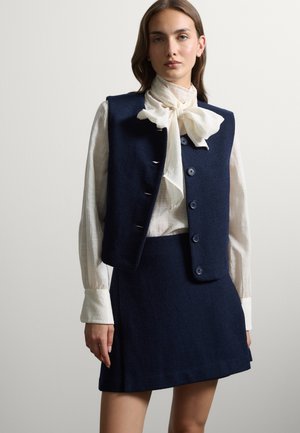 Donna che indossa un gilet blu navy con bottoni e gonna con una camicia bianca a maniche lunghe annodata con un fiocco al collo, in piedi su uno sfondo semplice.