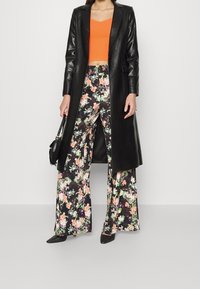 Schwarzer Lederlangmantel, orangefarbener Cropped-Top, florale weit geschnittene Hose. Enthält lebhafte Farben, Muster und strukturiertes Design. Schwarze spitze Absatzschuhe.