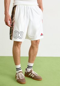 Shorts de sport blancs avec un panneau latéral camouflage, arborant le numéro noir "03" et le logo Adidas rouge. Texture lisse, ceinture élastique.