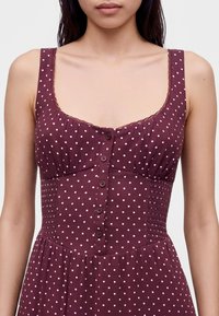 Femme portant une robe bordeaux sans manches à pois blancs, boutonnée sur le devant, avec un col carré et un bord en dentelle.