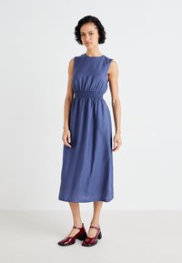 Blue sleeveless dress with gathered waist and knee-length hem; smooth texture. Paired with dark red chunky-heeled shoes.