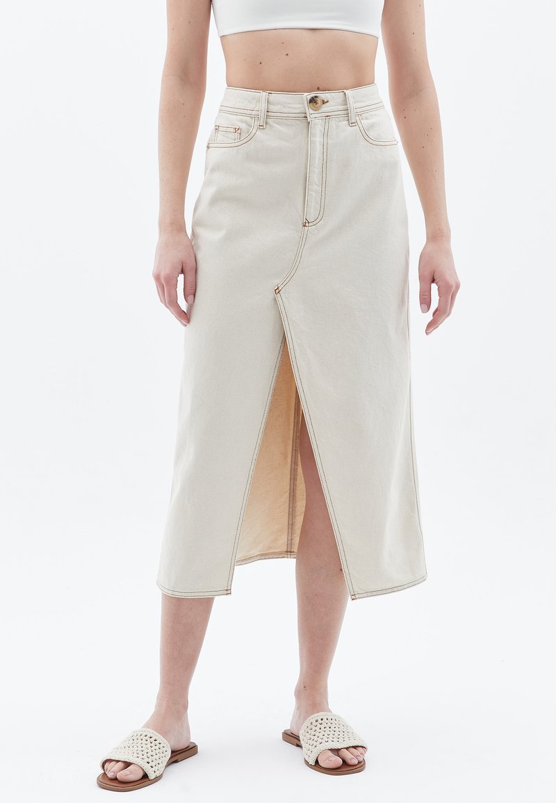 Beige denim midikjol med en framfals, sömnadsdetaljer och knappstängning i midjan. Modell bär avslappnade sandaler.