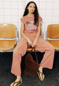 Haut ample rose avec des graphiques de palmiers, pantalon large coordonné, sac à main frangé marron et tongs à semelles jaunes. Accents matériels minimaux.