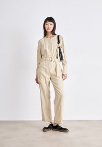Combinaison beige ceinturée avec un devant boutonné, des manches longues et une coupe évasée, associée à des baskets noires et un petit sac à main texturé.