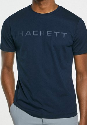 Mann trägt ein marineblaues kurzärmeliges T-Shirt mit dem Wort „HACKETT“ in etwas helleren blauen Buchstaben über der Brust.