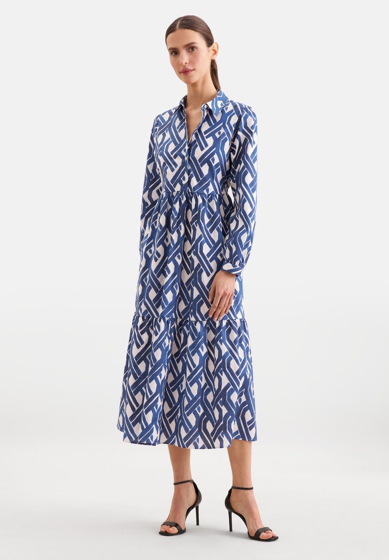 Femme portant une robe midi à manches longues boutonnée avec un motif bleu et blanc et des sandales noires à talons hauts avec une bride à la cheville, debout.