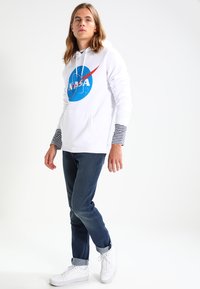 Fehér kapucnis pulóver NASA logóval, kék körrel és piros ívvel; csíkos hosszú ujjak alatta. Sötét farmernadrággal és fehér sportcipővel párosítva.