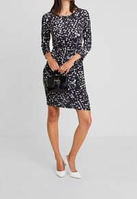 Robe à motifs noir et blanc avec des manches longues et une silhouette ajustée, associée à un petit sac à main noir et des chaussures à talons blancs.