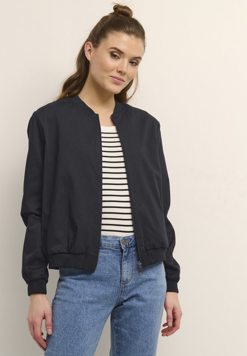 Marineblauer Bomberjacke mit rundem Kragen und gerippten Manschetten, kombiniert mit einem gestreiften Hemd und hochgeschnittenen blauen Jeans. Glatte Stoffstruktur.