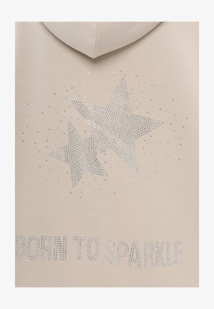Beige capuchontrui met zilveren rhinestones die in stervormen zijn gerangschikt, met daaronder de tekst "BORN TO SPARKLE" in rhinestones.