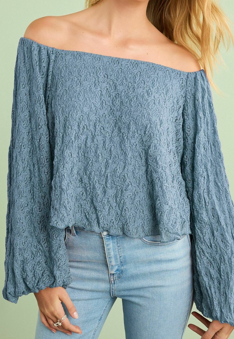 Femme portant un chemisier bleu texturé à manches longues et épaules dégagées, associé à un jean bleu clair, sur un fond vert clair.