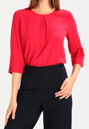 Femme portant un blouse rouge à manches longues avec des détails plissés, associée à un pantalon noir taille haute, posant avec une main sur la hanche.