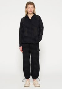 Zwarte overhemd met knoopsluiting en lasersnedes, gecombineerd met zwarte broek met trekkoord, voorzien van een cargozak en gouden hardwareaccenten.