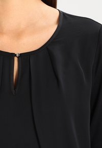Blusa nera con scollatura a chiave, dettagli plissettati e texture simile al satin. Chiusura con un piccolo bottone di metallo al collo.