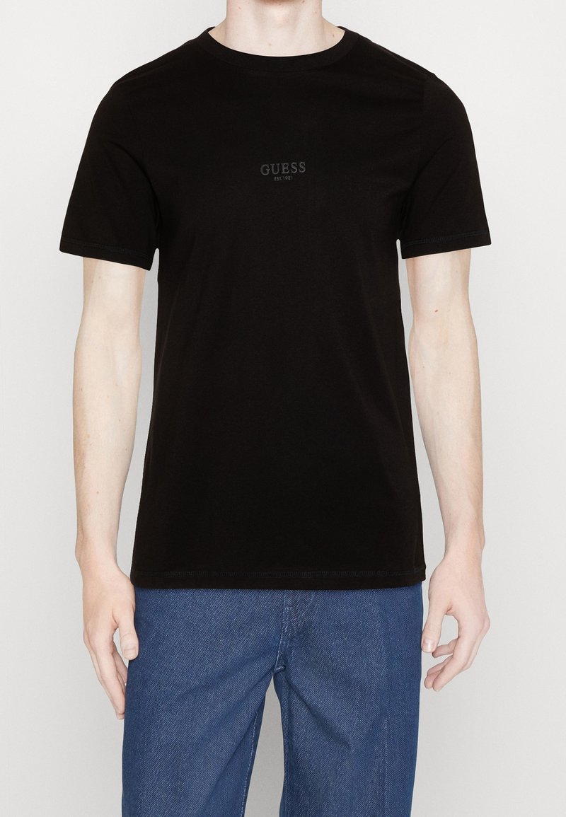 Homme portant un T-shirt noir à manches courtes uni avec le logo "GUESS EST. 1981" et un jean bleu, posant devant un fond blanc.