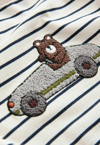 Svītraina kokvilnas auduma ar izšūtu lāci uz teksturēta транспорта, ar pelēkiem, zaļiem un oranžiem akcentiem uz gaiša fona.