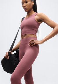 Γυναίκα που φοράει αμάνικο μωβ crop top και κολάν χωρίς ραφές, κρατώντας μια μαύρη τσάντα γυμναστηρίου με ιμάντα ώμου, μπροστά σε απλό λευκό φόντο.