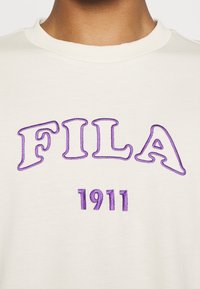 Sweatshirt en coton blanc cassé avec logo "FILA" brodé en violet et "1911" en dessous dans une police audacieuse et stylisée. Conception à col rond.