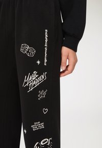 Zbliżenie czarnych spodni z białym tekstem w stylu ręcznego pisma i rysunkami, w tym kości, serca oraz hasłami takimi jak "Magia się zdarza" i "Przestań przepraszać za swoje emocje."