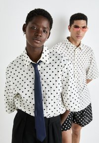 Biele košele s čiernymi bodkami, jedna s námorníckou kravatou. Pozadie je neutrálné; model vpravo nosí ružové slnečné okuliare a šortky.