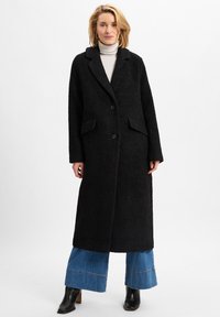 Schwarzer langer Mantel mit strukturierter Oberfläche, Reverskragen, zwei vorderen Taschen und zwei Knöpfen, getragen über weit geschnittenen blauen Jeans und Stiefeletten mit Absatz.