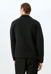 Sudadera negra con cuello clásico y puños acanalados, presenta una textura suave y una silueta ajustada. Vista posterior mostrada.