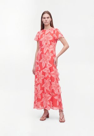 Mujer de pie vestida con un vestido largo color coral rosa con un gran estampado floral rosa claro y mangas cortas, combinado con sandalias de tacón coral.
