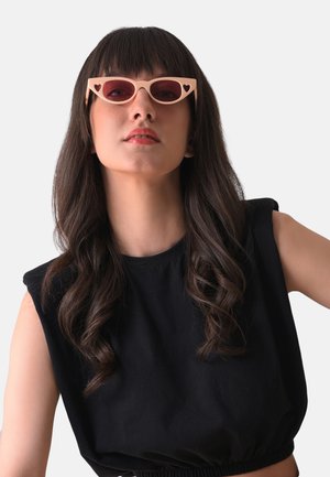 Occhiali da sole a forma di occhio di gatto con una montatura rosa chiaro e lenti rosa scuro, caratterizzati da ritagli a forma di cuore agli angoli. Indossati da una modella.