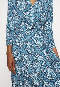 Femme portant une robe portefeuille à motif floral bleu et blanc avec des manches trois-quarts, tenant une partie de la jupe de la main droite.