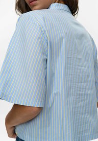 Korte mouwen shirt van lichtblauwe stof met gele en witte verticale strepen. Heeft een kraag en een relaxte pasvorm. Zachte textuur.