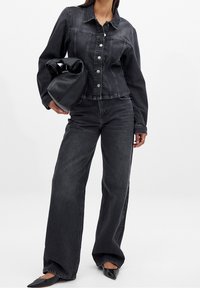 Veste en denim noir avec boutons argentés, associée à un jean noir à jambes larges. Sac hobo noir à la main, chaussures pointues noires. Fond uni.