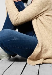 Beżowy, teksturowany kardigan zarzucony na nogi, w połączeniu z ciemnoniebieskimi dżinsami i białymi trampkami, siedzący na drewnianej podłodze.