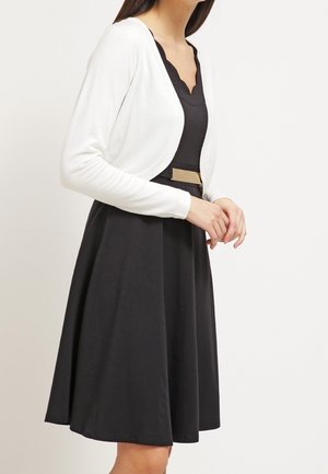Femme portant une robe noire avec une ceinture dorée et un cardigan blanc court à manches longues, debout devant un fond uni.