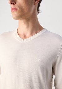 Maglione in maglia beige chiaro con scollo a V, colletto a coste e discreto logo del marchio ricamato sul petto. Texture liscia e leggero.