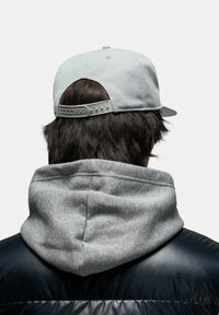Gorra de béisbol gris con correa ajustable y sudadera gris texturizada; posicionada contra una chaqueta acolchada negra, mostrando materiales en capas.