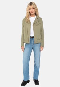 Giacca verde oliva con zip, colletto a rever e tasche laterali, abbinata a jeans dritti azzurri e stivali platform neri.