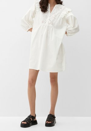 Femme portant une robe blanche ample avec des détails en dentelle ajourée et des manches bouffantes, associée à des sandales plateformes noires style pêcheur.