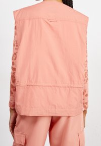 Person som bär en lös, ärmlös, ljusrosa väst över matchande cargobyxor och en långärmad topp med ärmar i rosa och bruna mönster.