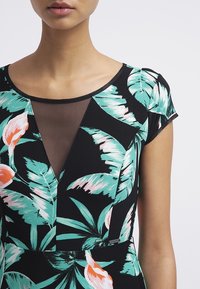 Kobieta w czarnej sukience z turkusowym i różowym wzorem tropikalnych liści i kwiatów, z przezroczystym wstawkiem z siateczki przy dekolcie i z krótkimi rękawami.