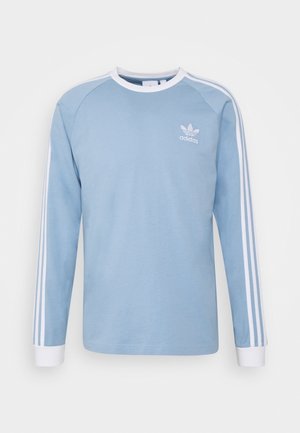 T-shirt à manches longues bleu avec des accents blancs. Il présente un col rond, trois rayures blanches sur chaque manche et un petit logo blanc sur la poitrine.