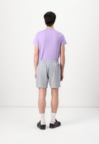 Ein männliches Model trägt ein lilafarbenes T-Shirt mit kurzen Ärmeln, graue gestreifte Shorts mit elastischem Bund und schwarze Schuhe mit weißen Socken.