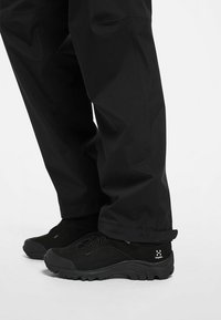 Black waterproof pants with relaxed fit, paired with black hiking shoes featuring textured suede and rubber soles. Subtle logo detail present.