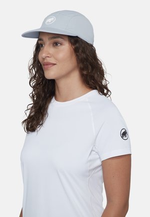 Vrouw met krullend haar, die een lichtblauwe pet en een wit shirt met korte mouwen draagt, met een zwart logo op de mouw.