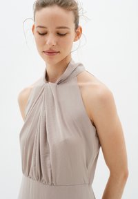 Helle taupe Halterkleid mit plissiertem Stoff, weicher Textur und tailliertem Schnitt. Verfügt über eine Nackenband und offene Schultern.