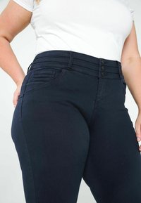 Jean bleu foncé avec une taille haute à triple bouton, un design à cinq poches et un tissu lisse et texturé.
