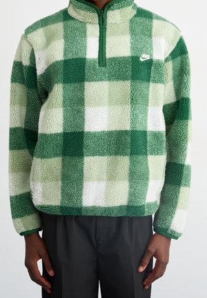 Person trägt einen grün-weiß karierten Fleece-Pullover mit Viertelreißverschluss und Nike-Logo auf der Brust, kombiniert mit schwarzen Hosen.