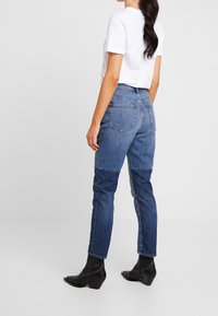 Jeans de mezclilla azul de talle alto con un diseño bicolor, que cuentan con bolsillos traseros y un corte recto, combinados con botas negras hasta el tobillo.
