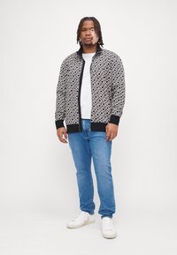 Veste bomber à motif noir et blanc texturée avec fermeture éclair à l'avant, portée sur une chemise blanche et assortie avec un jean bleu clair et des baskets blanches.