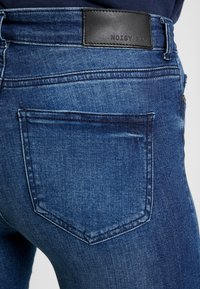 Pantalones de mezclilla con un lavado azul oscuro, que presentan un bolsillo trasero y una etiqueta de cuero cosida etiquetada "NOISY MAY". Textura suave, diseño ajustado.