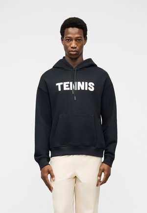 Jeune homme portant un sweat à capuche noir avec le texte "TENNIS" et un pantalon beige clair, debout devant un fond clair uni.